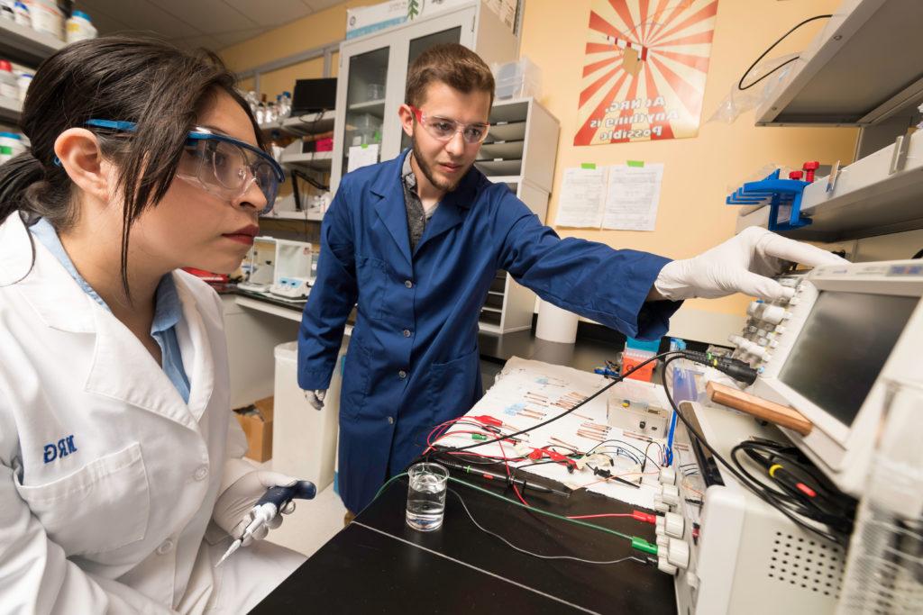 Two student researchers in a lab, wearing safety equipment and altering electronic panels